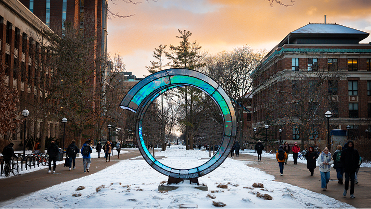 People walking on a snowy campus path near a large circular sculpture at sunset.