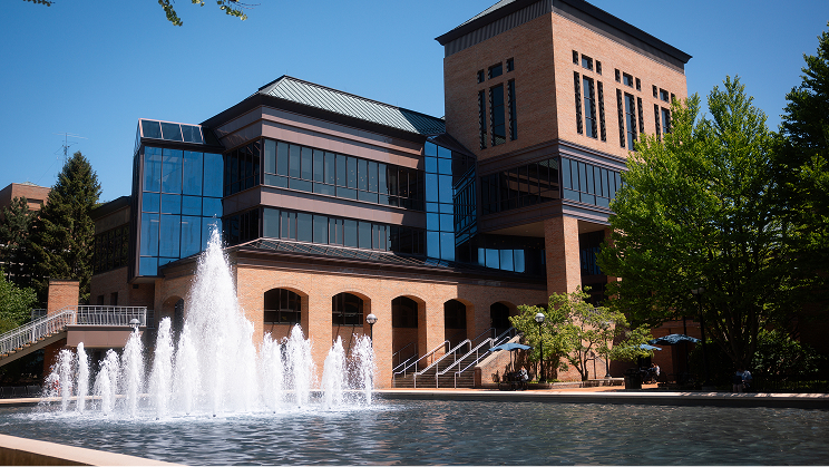 Modern brick building with large windows behind a fountain, surrounded by trees on a sunny day.